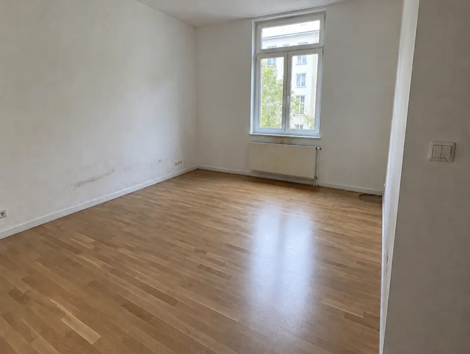 The same living room at move-out with scuffs on the wall, a stain under the window and a damaged floor patch